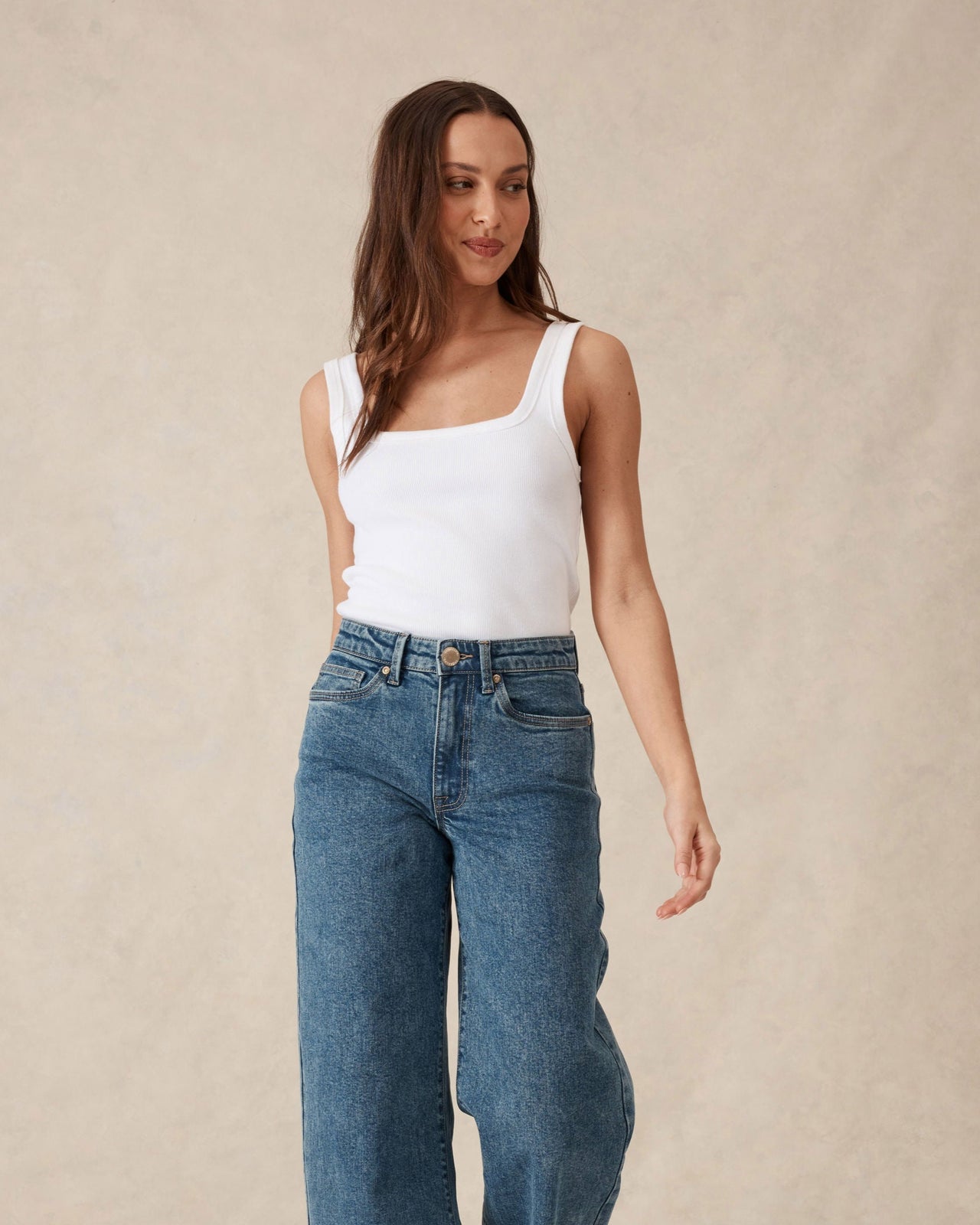 Woman wearing a white tank top and blue jeans against a beige background