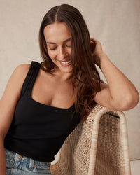 Thumbnail for Woman sitting on a wicker chair wearing a black tank top and blue jeans, smiling.