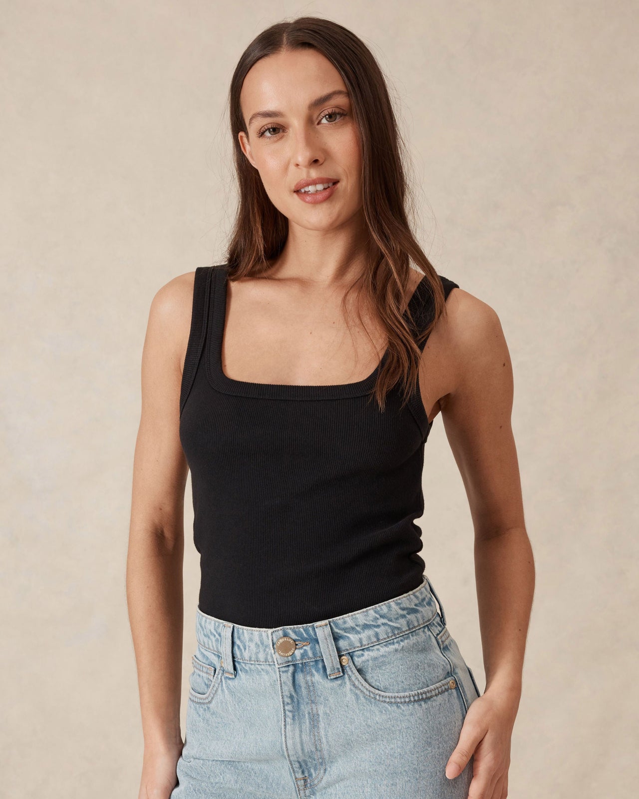 Woman wearing a black tank top and light blue jeans against a beige background