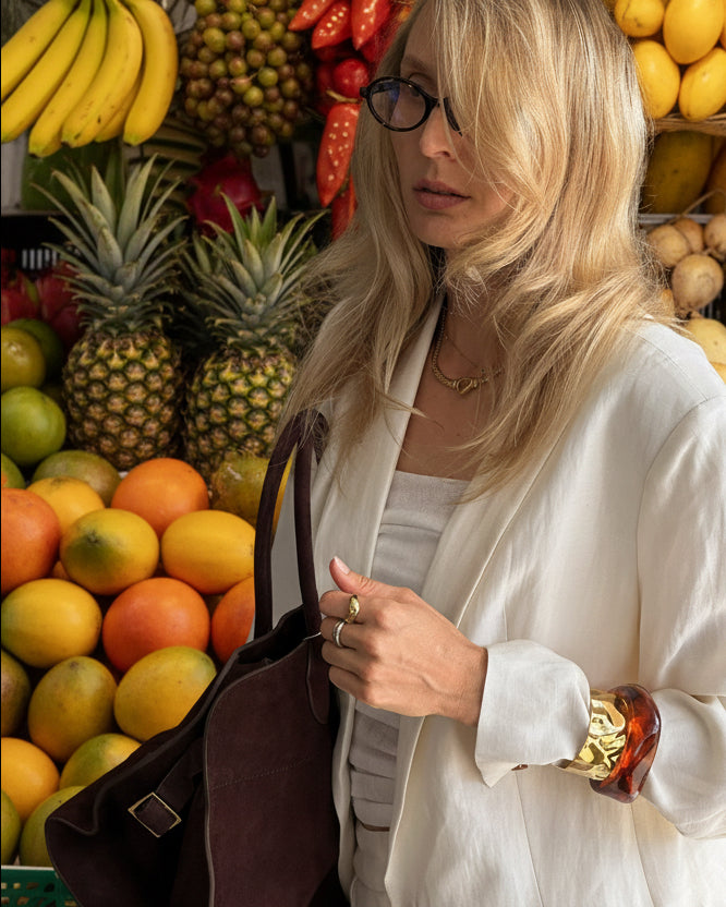 Woman with blonde hair wearing glasses, holding a brown bag against a dark background