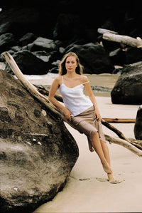 Thumbnail for Woman wearing white silk camisole and taupe silk slip skirt leaning on a large rock at the beach