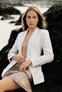 Thumbnail for Model wearing a white linen blazer and taupe silk slip skirt posing on a rock by the beach