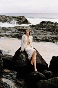 Thumbnail for Model wearing white linen blazer and cream silk slip skirt posing on a rock by the beach