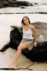 Thumbnail for Woman wearing white silk camisole and taupe silk slip skirt sitting on a rock by the beach
